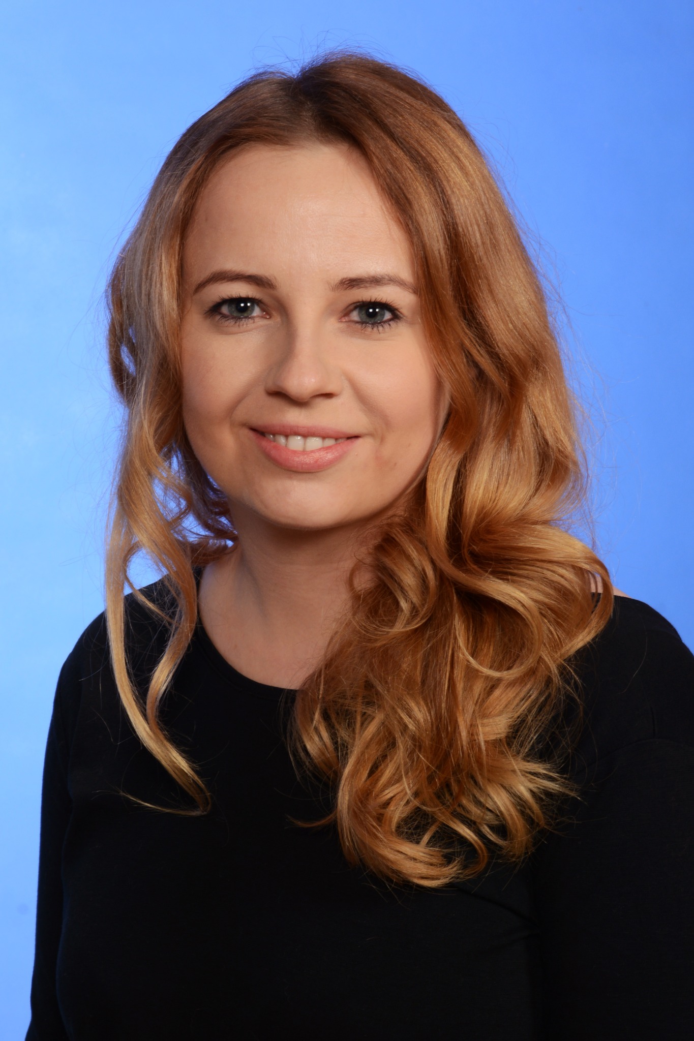 Portret uśmiechniętej kobiety o długich, rudych włosach na jednolitym, błękitnym tle.