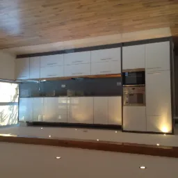 Nowoczesna kuchnia z białymi szafkami z połyskiem, drewnianym sufitem i listwą podłogową z punktowym oświetleniem LED.