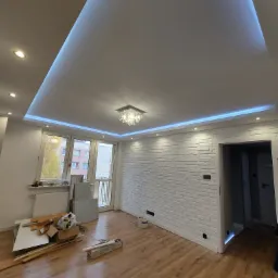 Pokój po remoncie z drewnianą podłogą, ścianą z białej cegły, podwieszanym sufitem z niebieskim oświetleniem LED i kryształowym żyrandolem, widoczne ślady prac remontowych.