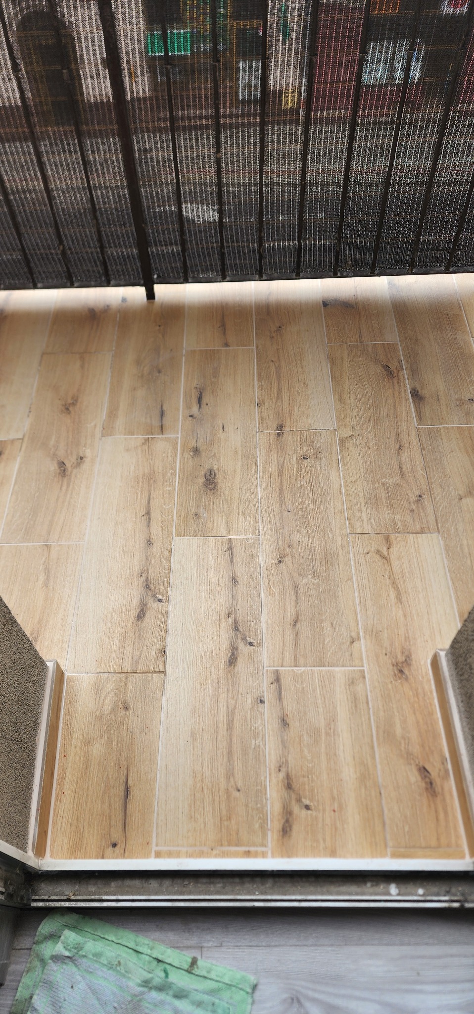 Balkon wyłożony płytkami imitującymi drewno, widok z góry spod czarnej balustrady. Jasne, podłużne płytki ułożone równolegle, z widocznymi fugami.