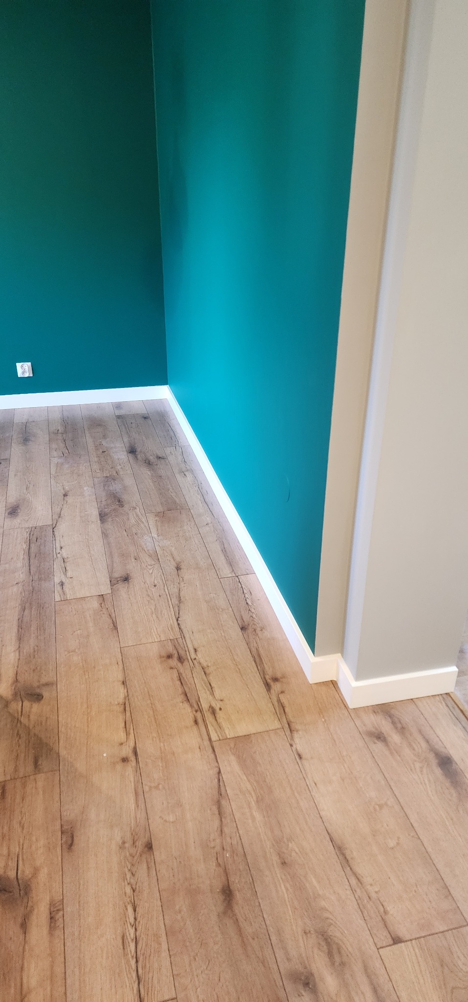 Ukończone wnętrze: drewnopodobne panele, białe listwy przypodłogowe i ściany w odcieniach turkusu i szarości. Widoczny kontakt elektryczny.