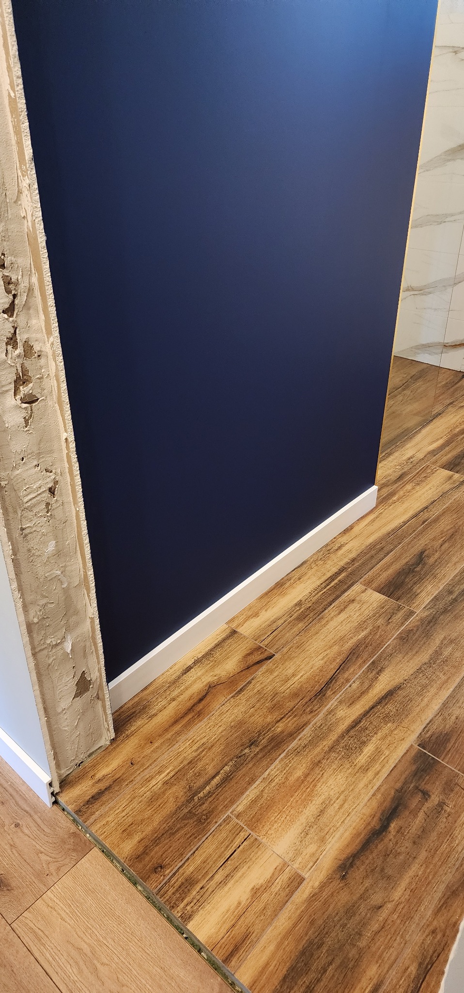 Fragment ściany w kolorze granatowym z białą listwą przypodłogową i drewnopodobną podłogą z płytek. Wykończenie w trakcie prac, widoczne surowe obrzeże.