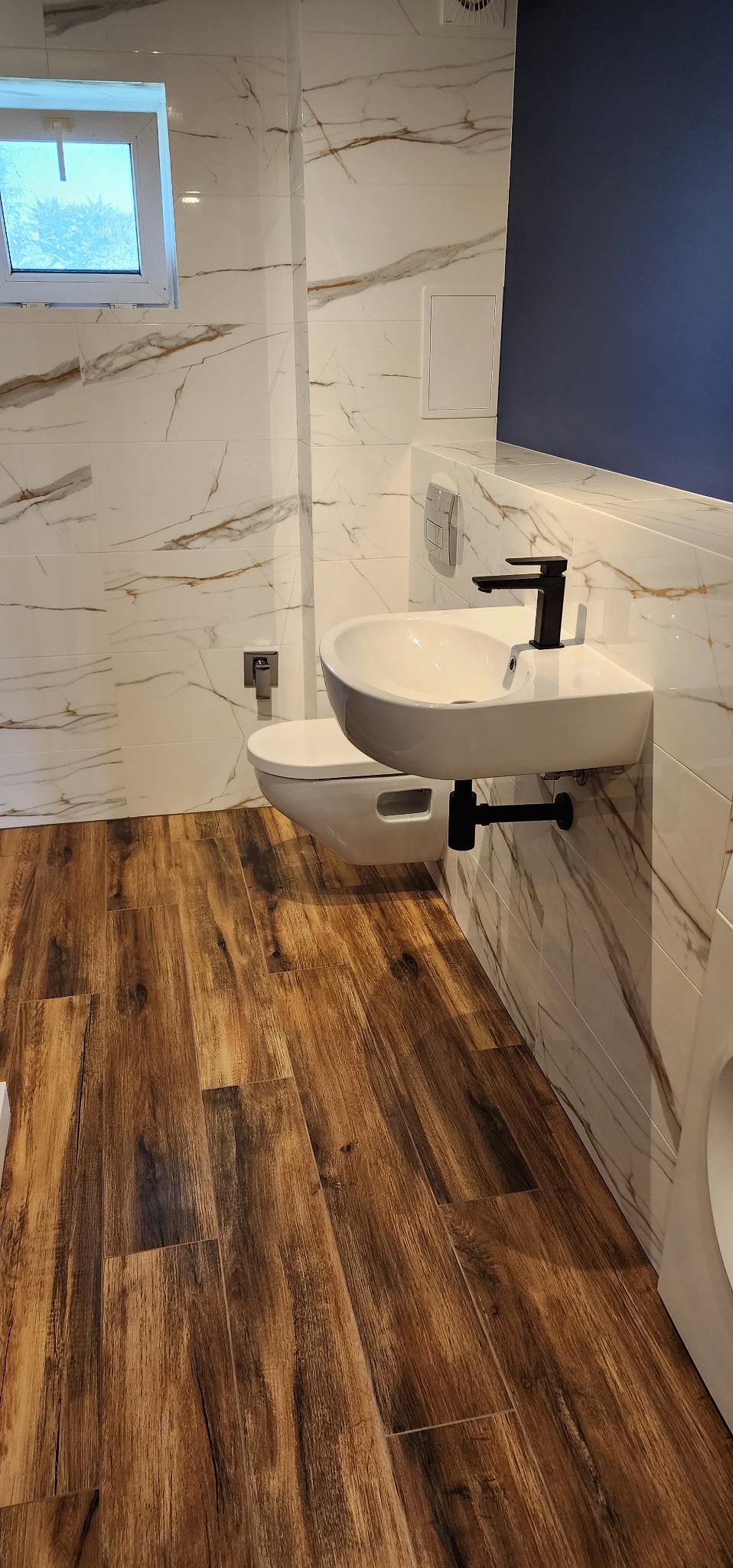 Nowoczesna łazienka po remoncie: białe płytki imitujące marmur na ścianach, drewnopodobne panele na podłodze, wisząca umywalka z czarną armaturą i toaleta.