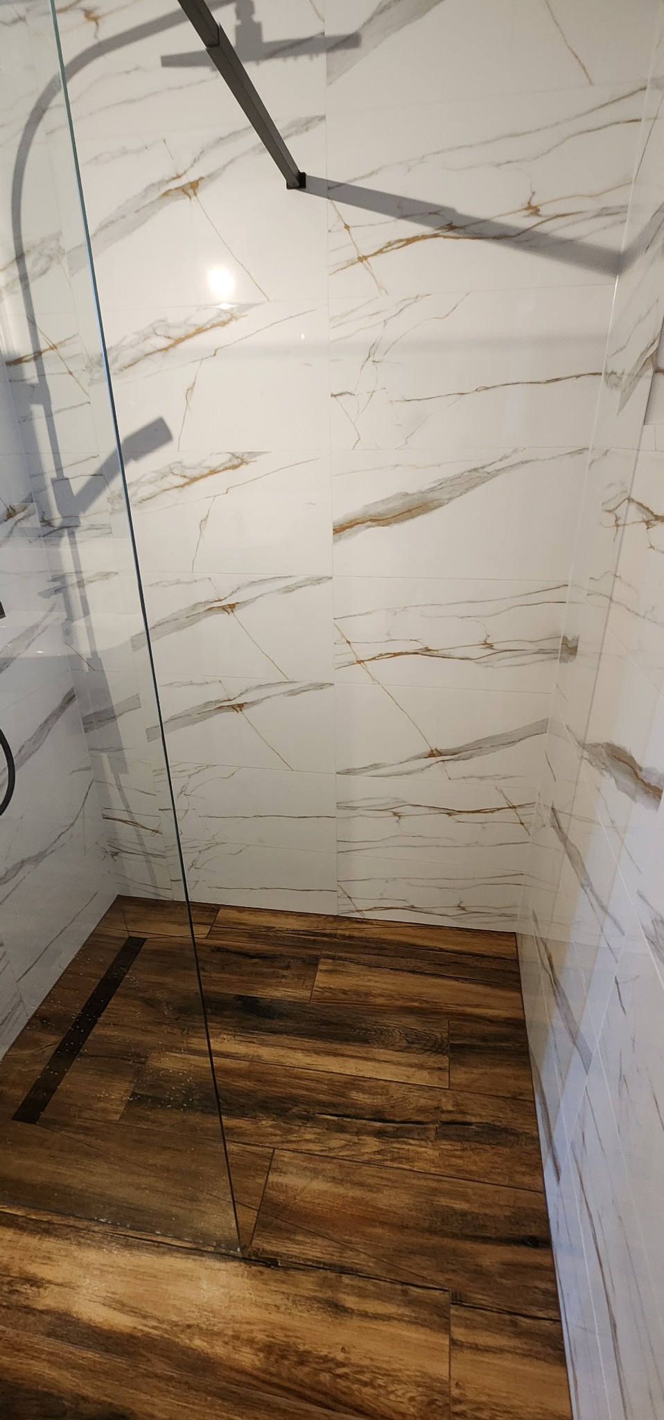 Kabina prysznicowa z drewnopodobną podłogą i marmurowymi płytkami ściennymi z złotymi żyłami. Widoczny odpływ liniowy.