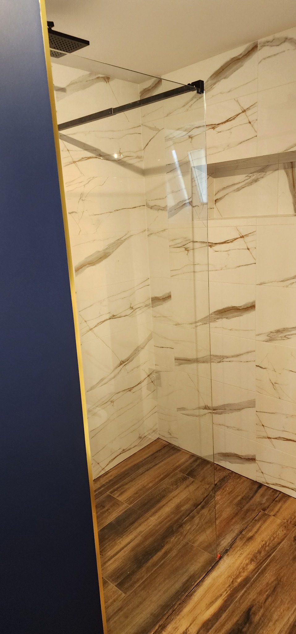 Nowoczesna kabina prysznicowa ze szklaną ścianką, wykończona płytkami imitującymi marmur i drewnopodobną podłogą. Widoczny czarny panel natryskowy.