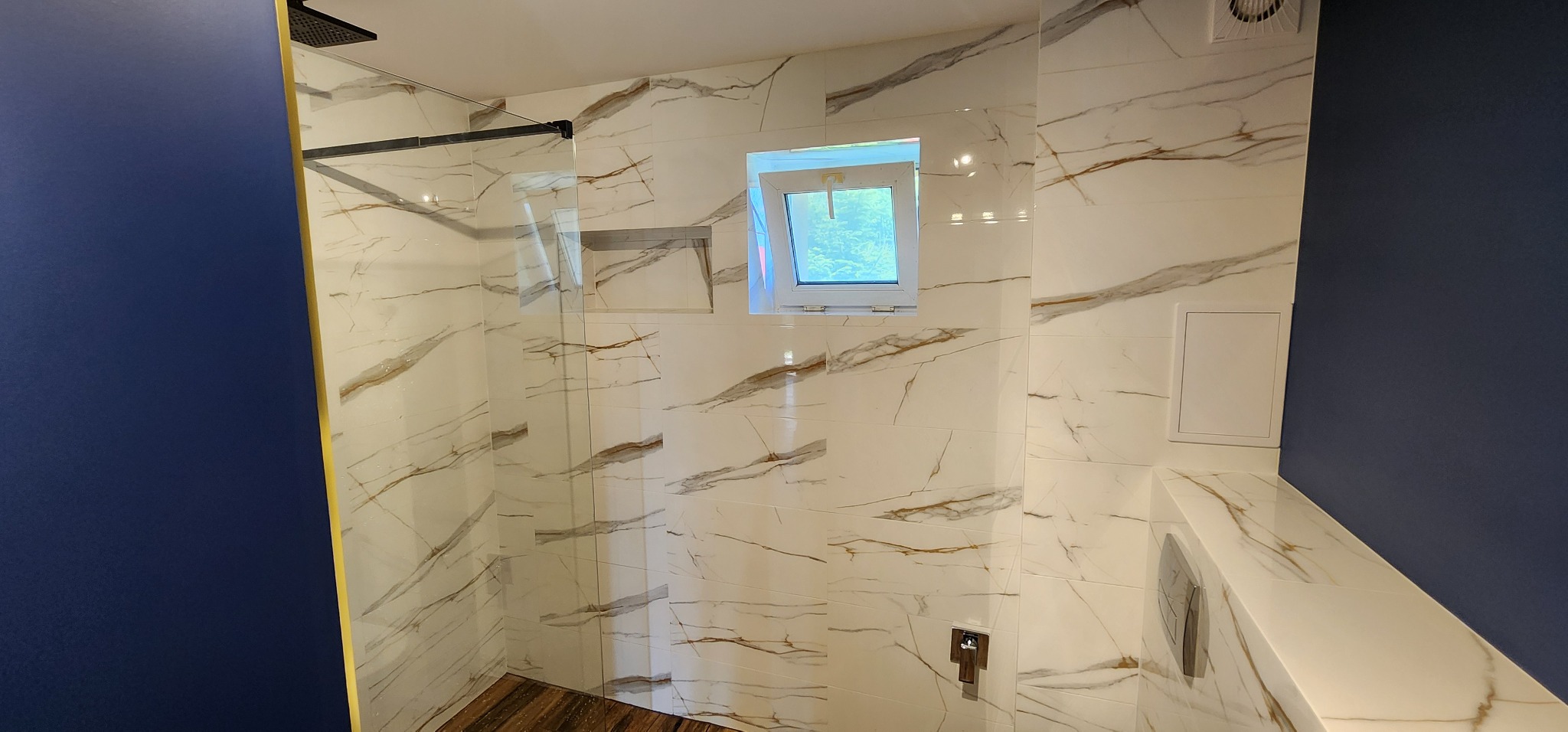 Białe płytki marmurowe z brązowymi żyłami w kabinie prysznicowej z czarną ramą i deszczownicą. Małe okno z widokiem na zieleń. Ściana pomalowana na granatowo.