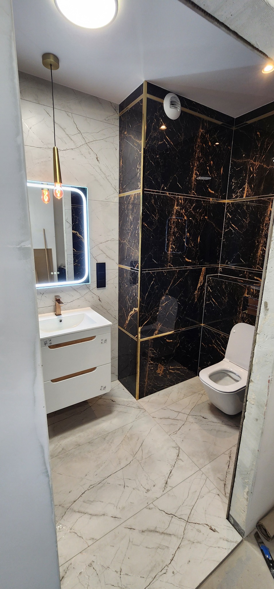 Nowoczesna łazienka z marmurowymi płytkami w odcieniach bieli i czerni, złotymi akcentami, umywalką z szafką i podświetlanym lustrem. Widoczna toaleta i wentylator.