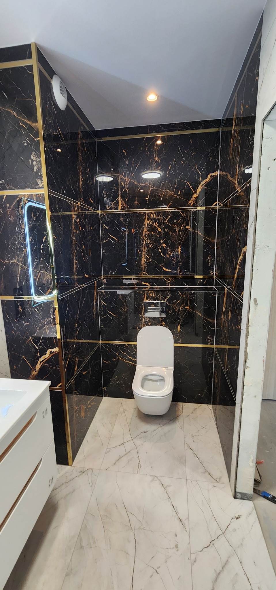 Elegancka łazienka z czarnymi płytkami imitującymi marmur, złotymi akcentami i podświetlanym lustrem. Biała toaleta i umywalka dopełniają nowoczesny wystrój.