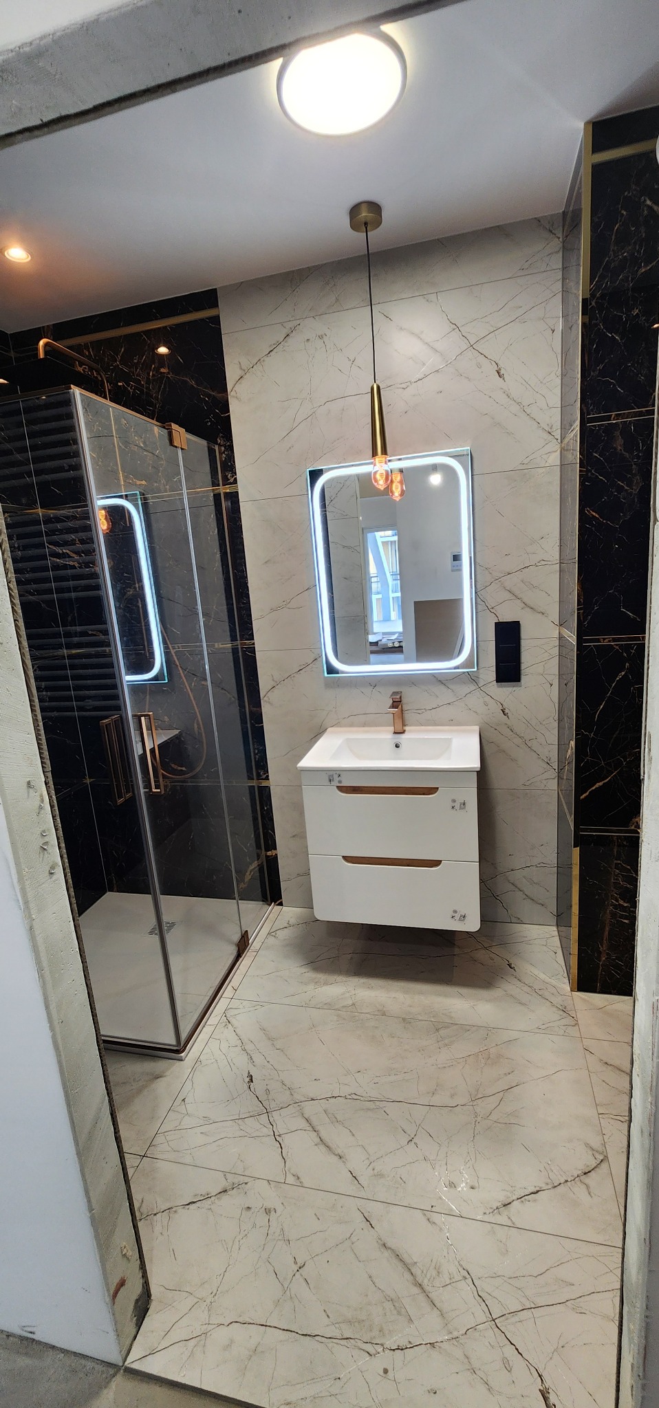 Nowoczesna łazienka z kabiną prysznicową, umywalką z szafką i podświetlanym lustrem. Marmurowe płytki na podłodze i ścianach, złote akcenty.