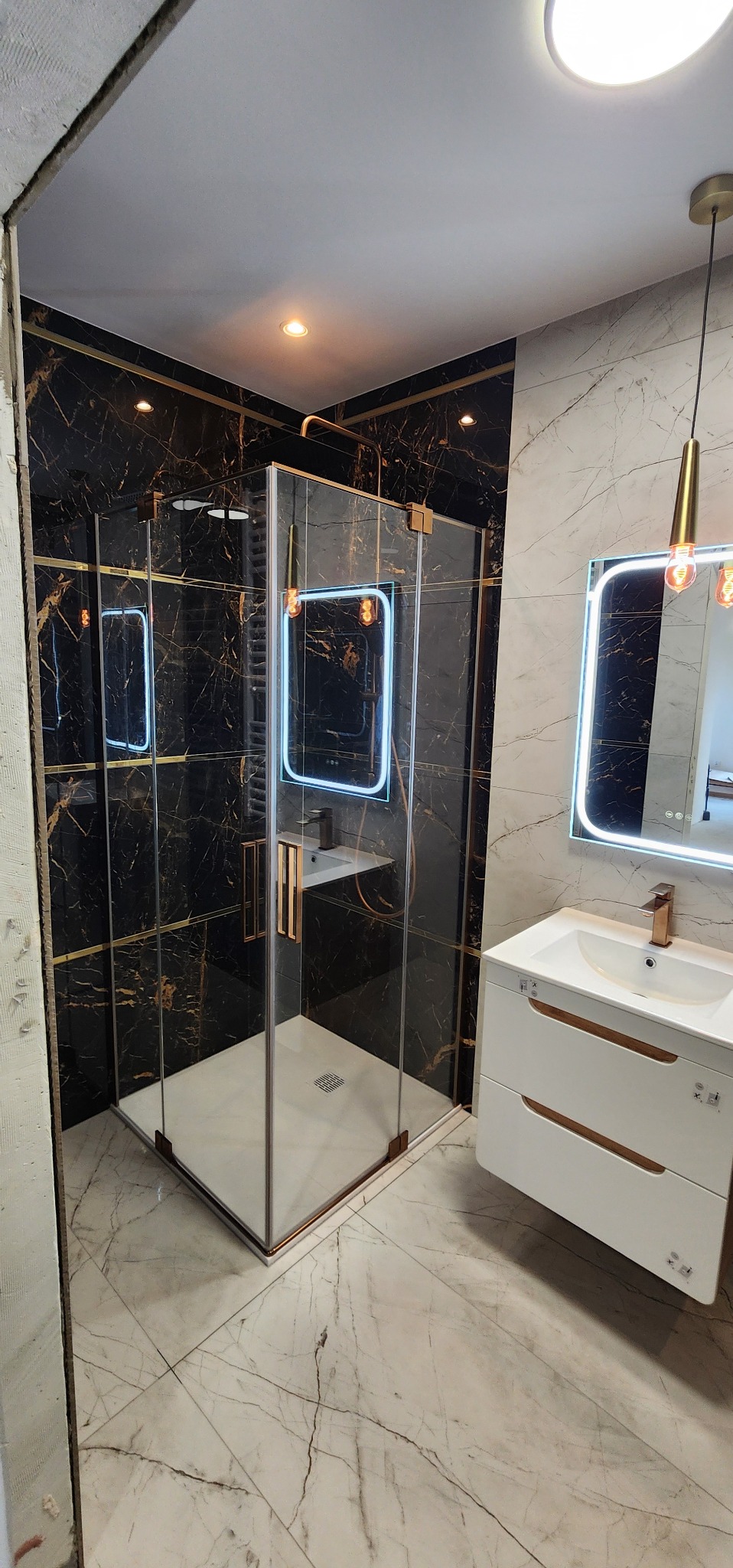 Nowoczesna kabina prysznicowa z czarnymi płytkami imitującymi marmur i złotymi akcentami, umywalka z szafką i lustro z podświetleniem LED w wykończonej łazience.