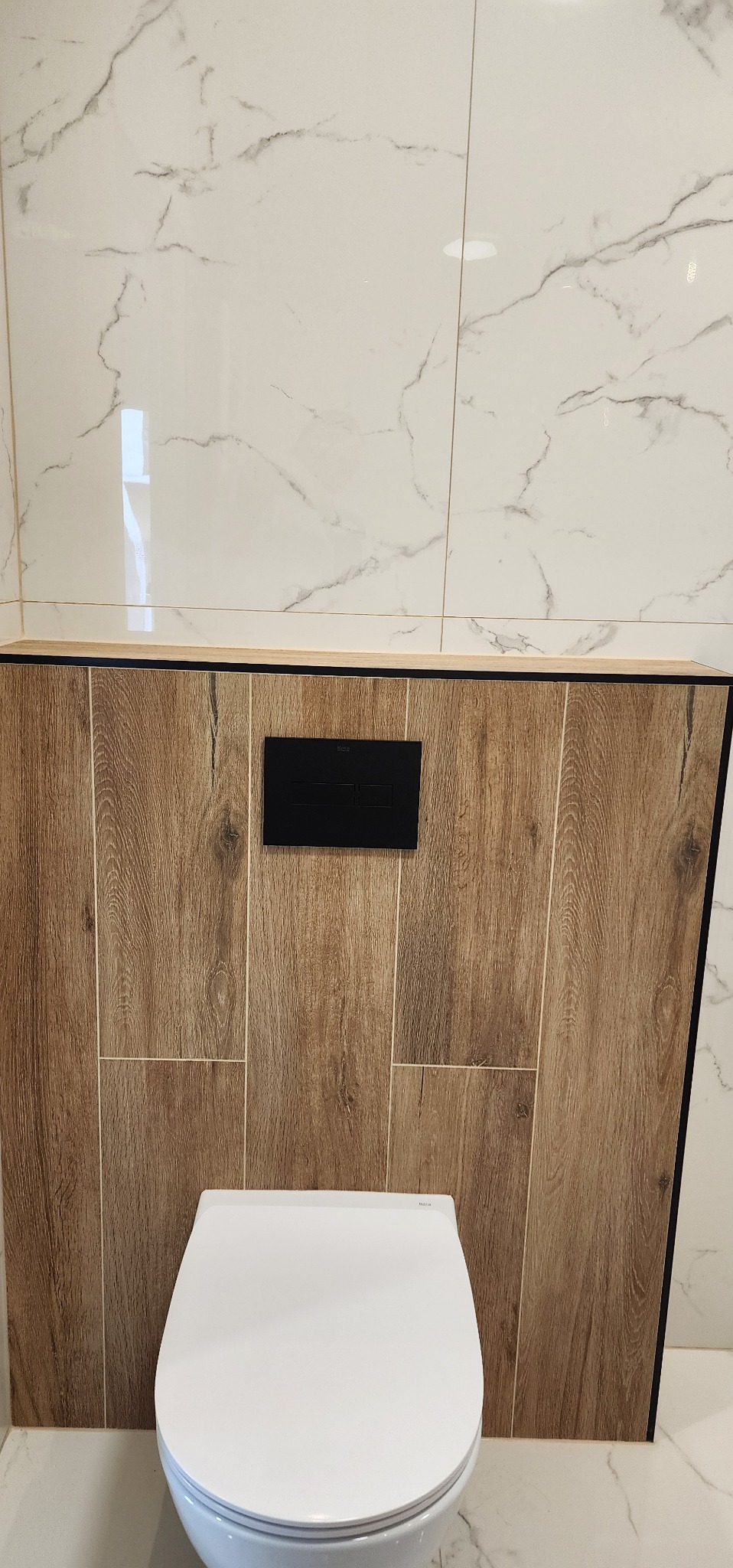 Nowoczesna toaleta podwieszana z czarnym przyciskiem spłukującym, obudowana płytkami imitującymi drewno, na tle białych płytek marmurowych. Minimalistyczny design łazienki.