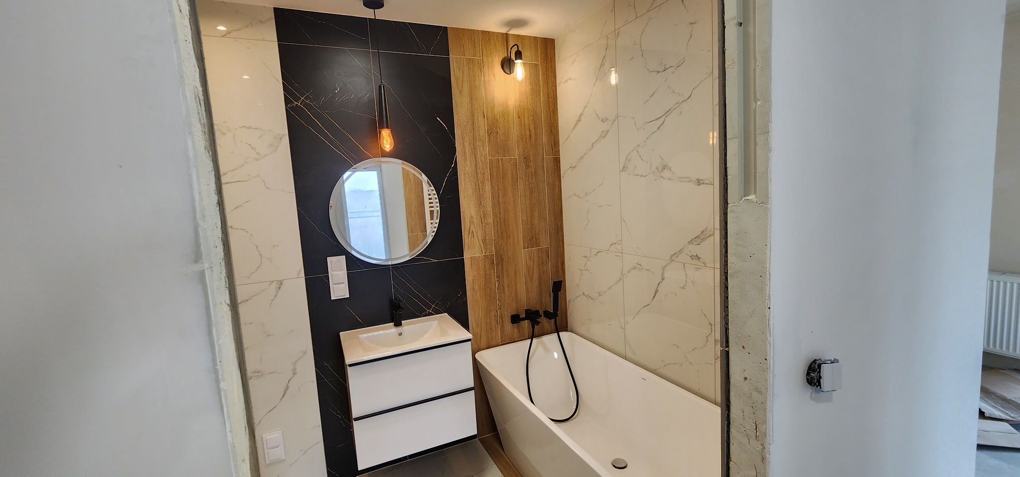 Nowoczesna łazienka z wanną, umywalką z szafką i okrągłym lustrem. Ściany wyłożone płytkami imitującymi marmur i drewno, oświetlenie punktowe i wisząca lampa.