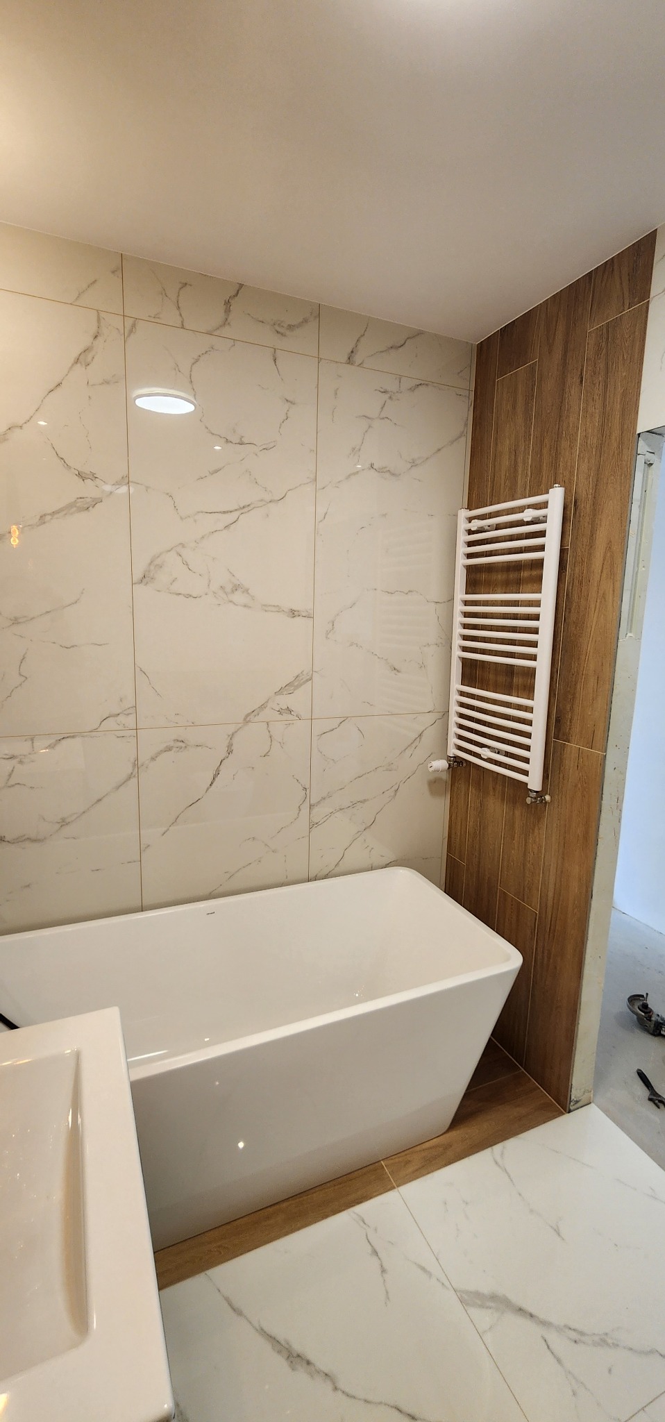 Nowoczesna łazienka z wanną, grzejnikiem drabinkowym i marmurowymi płytkami. Widoczne detale wykończenia i oświetlenie sufitowe.
