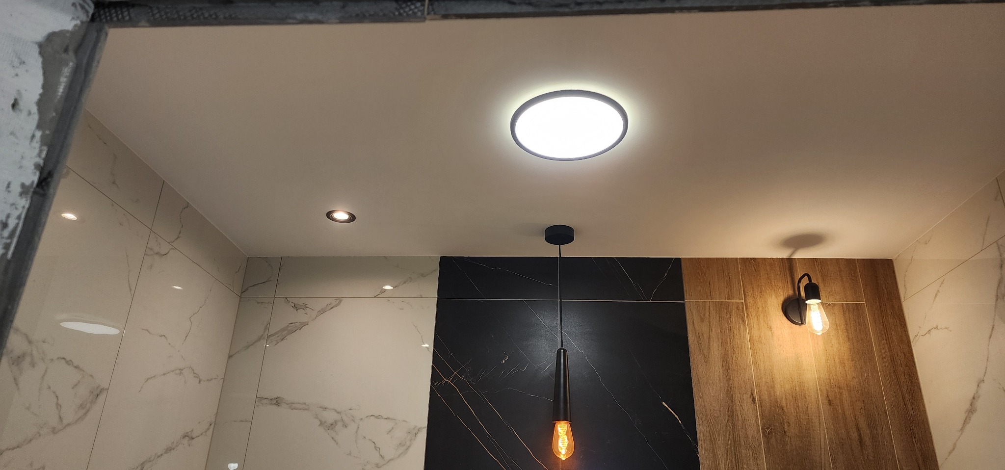 Nowoczesna łazienka z marmurowymi płytkami, oświetleniem sufitowym i dekoracyjną lampą wiszącą. Widoczne połączenie różnych wzorów płytek na ścianach.