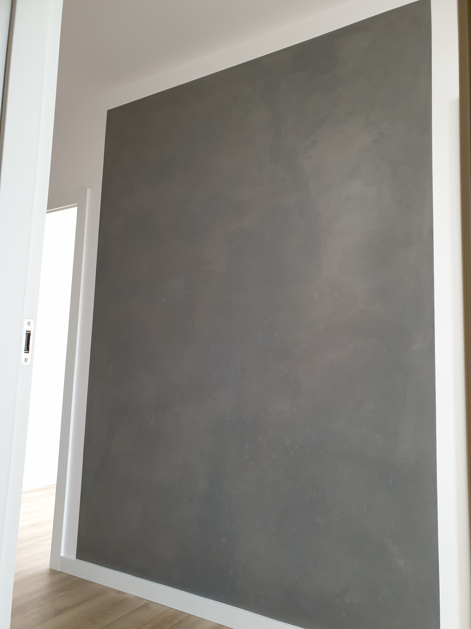 Szara ściana z efektem dekoracyjnym, widoczna z perspektywy otwartych białych drzwi, z fragmentem parkietu na podłodze.