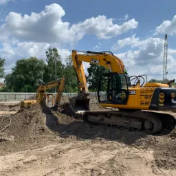 Dwie żółte koparki JCB na placu budowy, w tle widoczny dźwig i pracownik w kamizelce odblaskowej, błękitne niebo z obłokami.