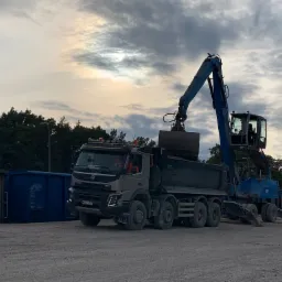 Ciężar&oacute;wka Volvo z naczepą wywrotką i niebieski żuraw załadowczy na placu, załadunek materiału do naczepy, widoczne kontenery, pochmurne niebo.