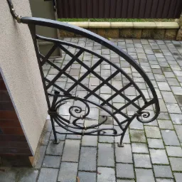 Metalowa, dekoracyjna konstrukcja nad schodami wejściowymi, z kręconymi elementami i kratownicą, zamontowana na ścianie obłożonej cegłą i tynkiem, z kostką brukową w tle.