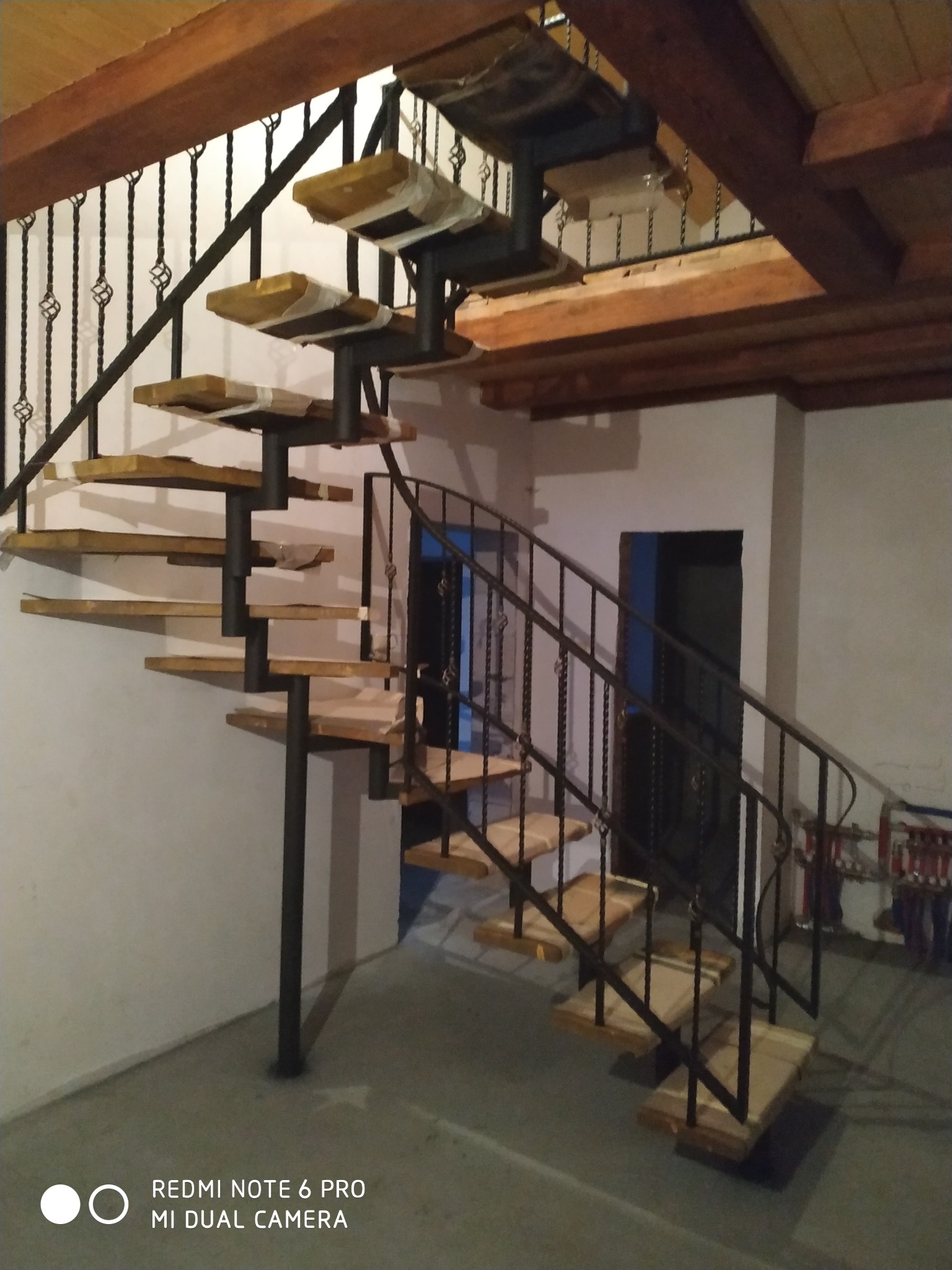Spiralne schody z drewnianymi stopniami i metalową balustradą w trakcie montażu, stopnie zabezpieczone folią, widoczne drewniane belki stropowe.