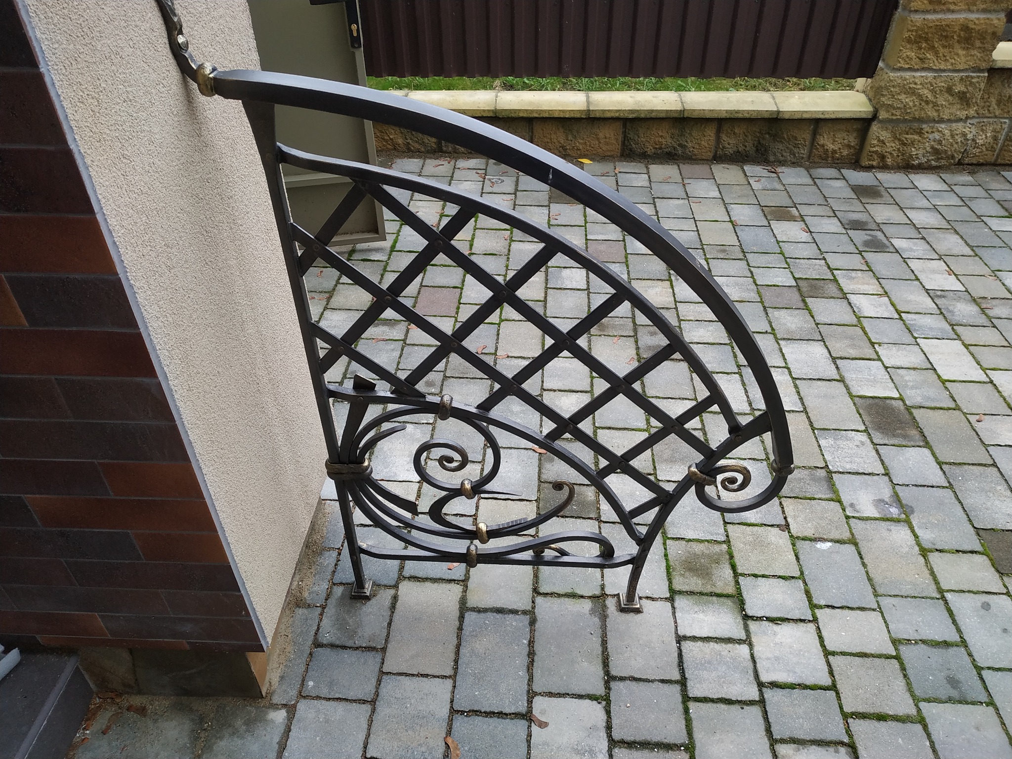 Metalowa, dekoracyjna konstrukcja nad schodami wejściowymi, z kręconymi elementami i kratownicą, zamontowana na ścianie obłożonej cegłą i tynkiem, z kostką brukową w tle.