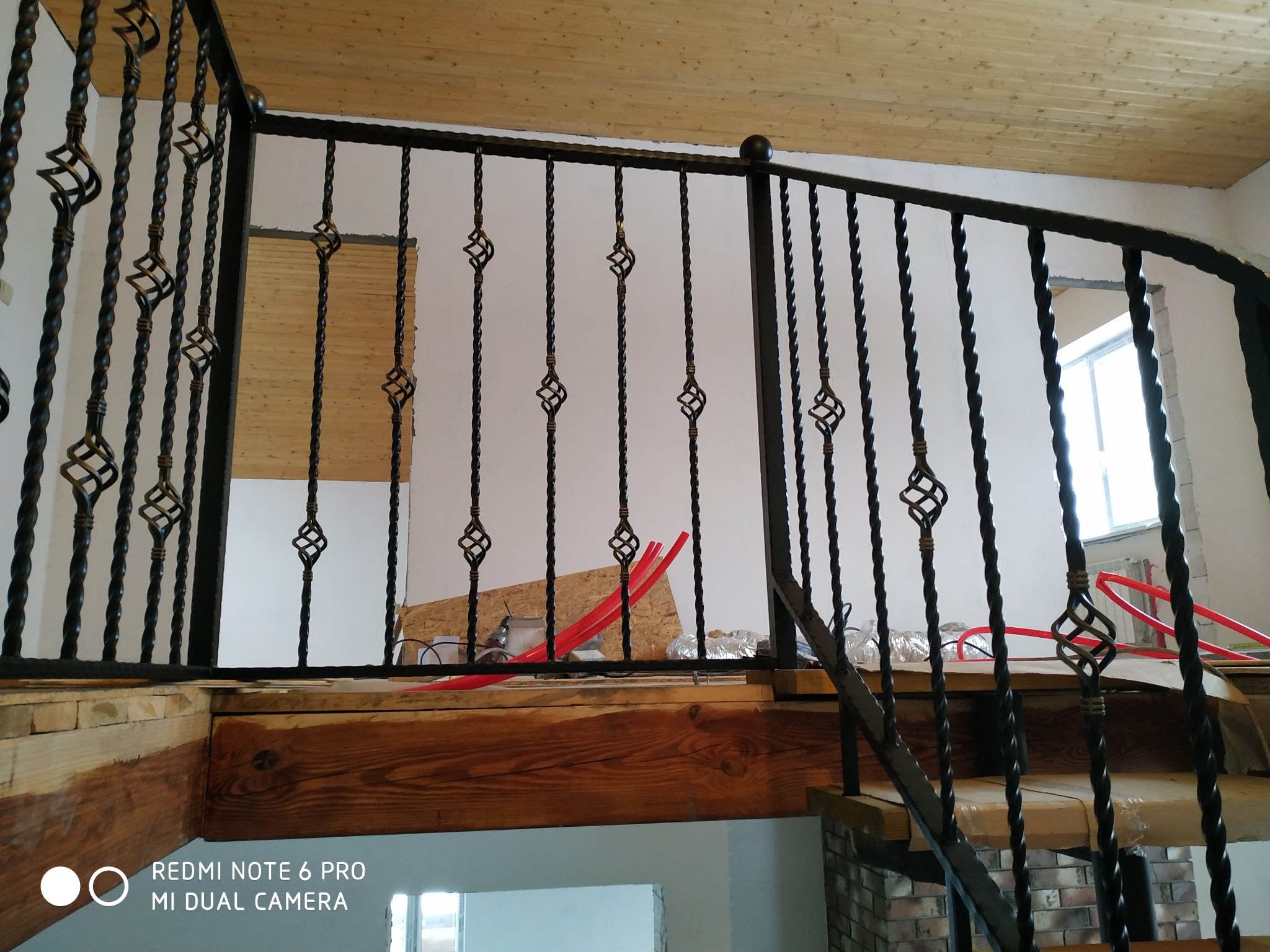 Metalowa balustrada schodowa z ozdobnymi, skręconymi elementami, widok z dołu na drewniany strop i fragment schodów w trakcie montażu instalacji grzewczej z czerwonymi rurami.