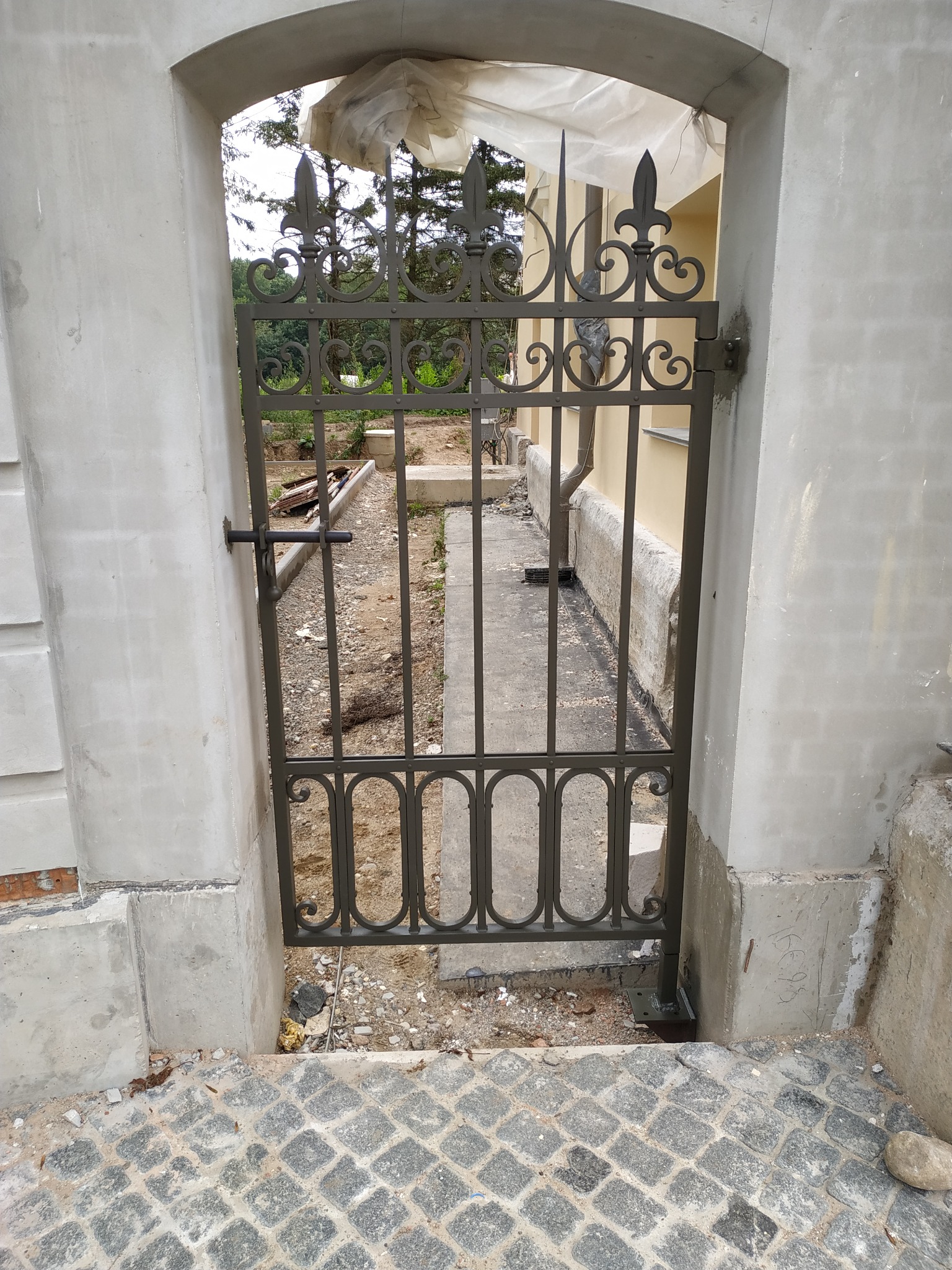 Metalowa furtka z ozdobnymi elementami w kształcie lilii, osadzona w surowym, betonowym łuku wejściowym, widoczna z perspektywy brukowanego podjazdu.