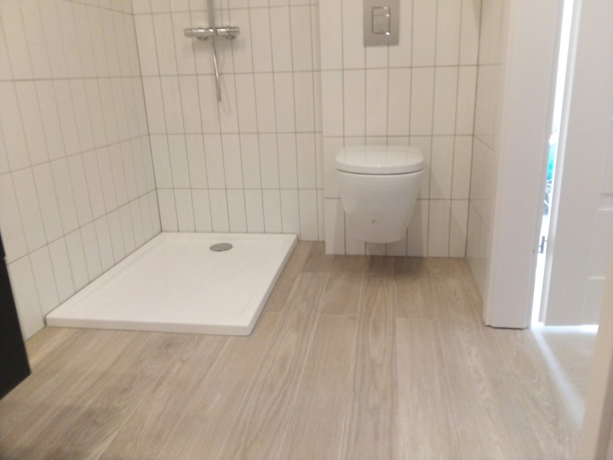 Nowoczesna łazienka z białymi płytkami, kabiną prysznicową bez brodzika i podwieszaną toaletą, wykończona panelami podłogowymi imitującymi drewno.