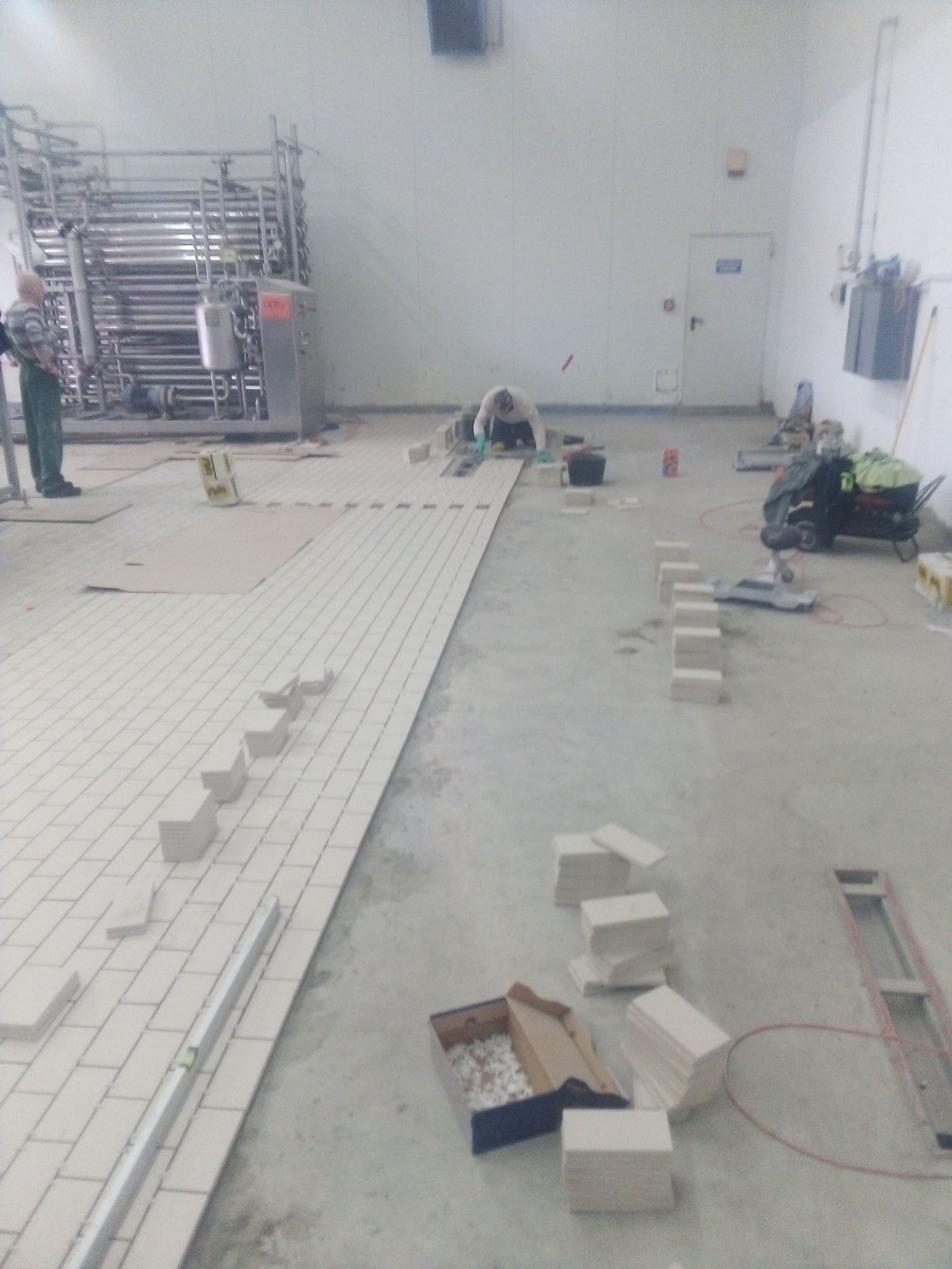 Układanie jasnych płytek podłogowych w pomieszczeniu przemysłowym, widoczni pracownicy w trakcie pracy, narzędzia i materiały budowlane.