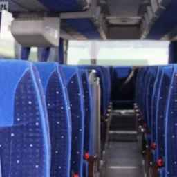 Wnętrze autobusu z rzędami niebieskich, tapicerowanych foteli z wzorem w kropki, widok wzdłuż korytarza w kierunku wyjścia.