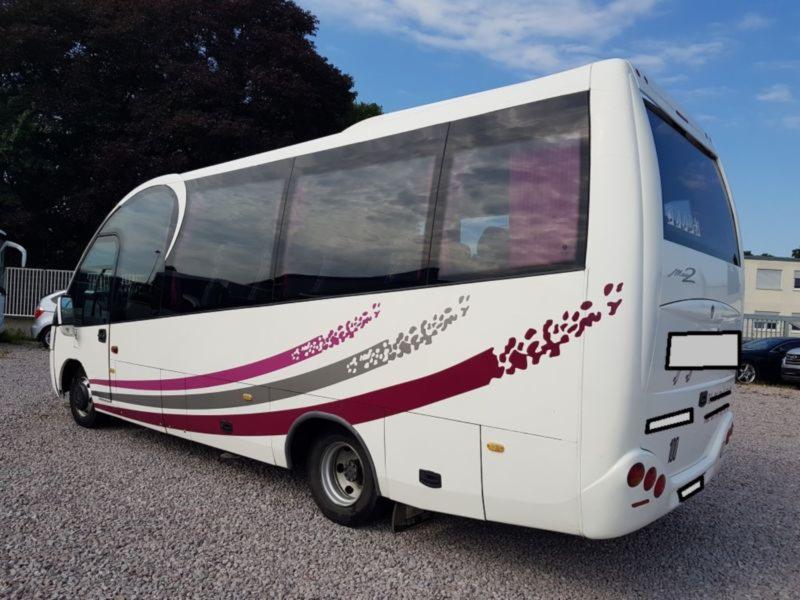 Biały bus z fioletowo-szarymi pasami i wzorami na karoserii, zaparkowany na żwirowym parkingu, widok z tyłu i boku.