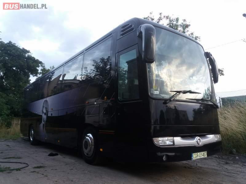 Czarny autokar z odbiciem chmur w szybach, zaparkowany na szutrowym podłożu, z widocznym zarysem twarzy na boku pojazdu.