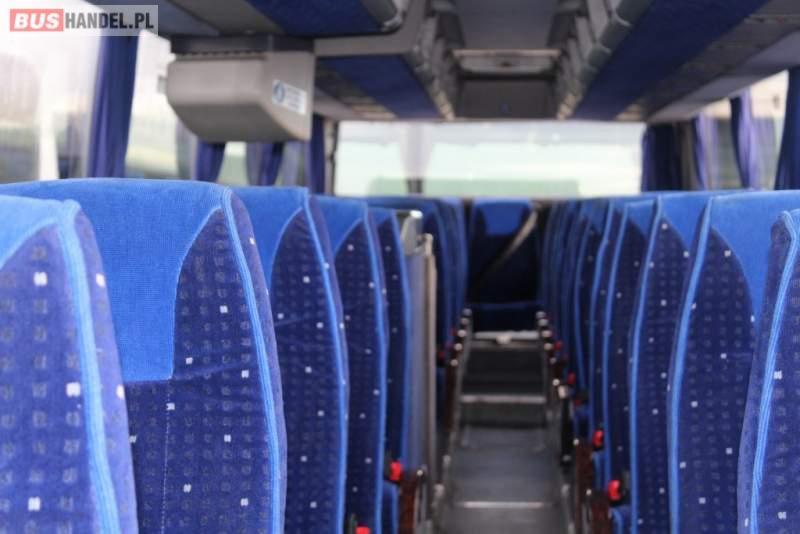 Wnętrze autobusu z rzędami niebieskich, tapicerowanych foteli z wzorem w kropki, widok wzdłuż korytarza w kierunku wyjścia.