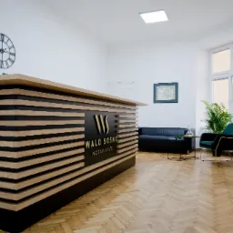 Nowoczesne wnętrze kancelarii notarialnej z recepcją z poziomych desek, czarną sofą, zielonym fotelem i parkietem w jodełkę.