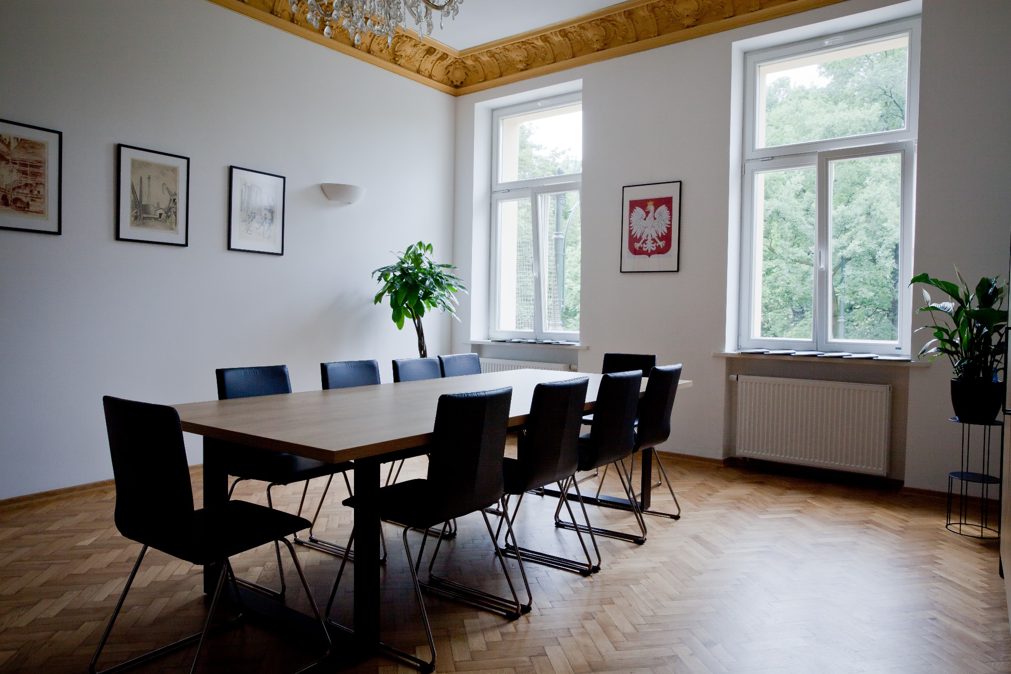 Sala konferencyjna w kancelarii notarialnej z drewnianym stołem, czarnymi krzesłami, parkietem w jodełkę oraz oknami z widokiem na zieleń, ozdobiona obrazami i godłem Polski.