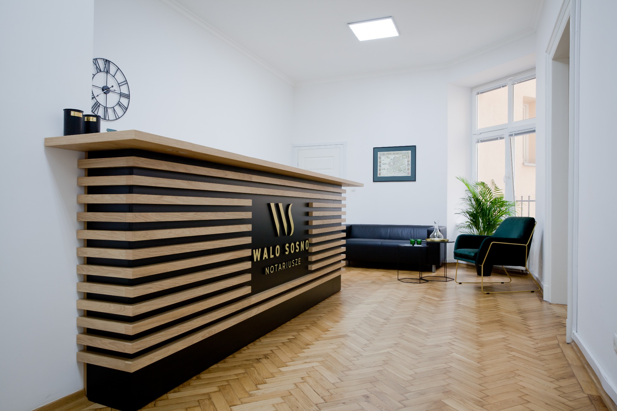 Nowoczesne wnętrze kancelarii notarialnej z recepcją z poziomych desek, czarną sofą, zielonym fotelem i parkietem w jodełkę.