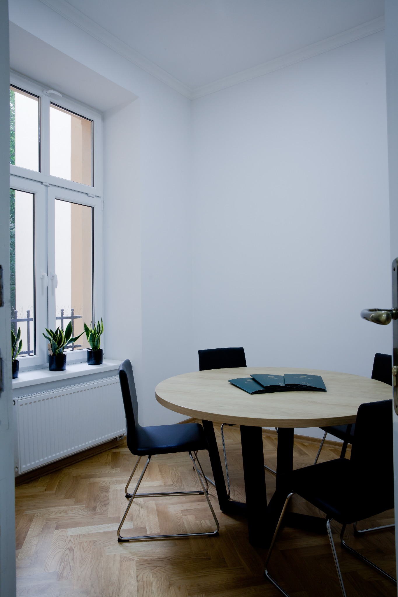Jasne biuro z okrągłym stołem, czarnymi krzesłami i trzema zielonymi teczkami na blacie, widok zza uchylonych drzwi.