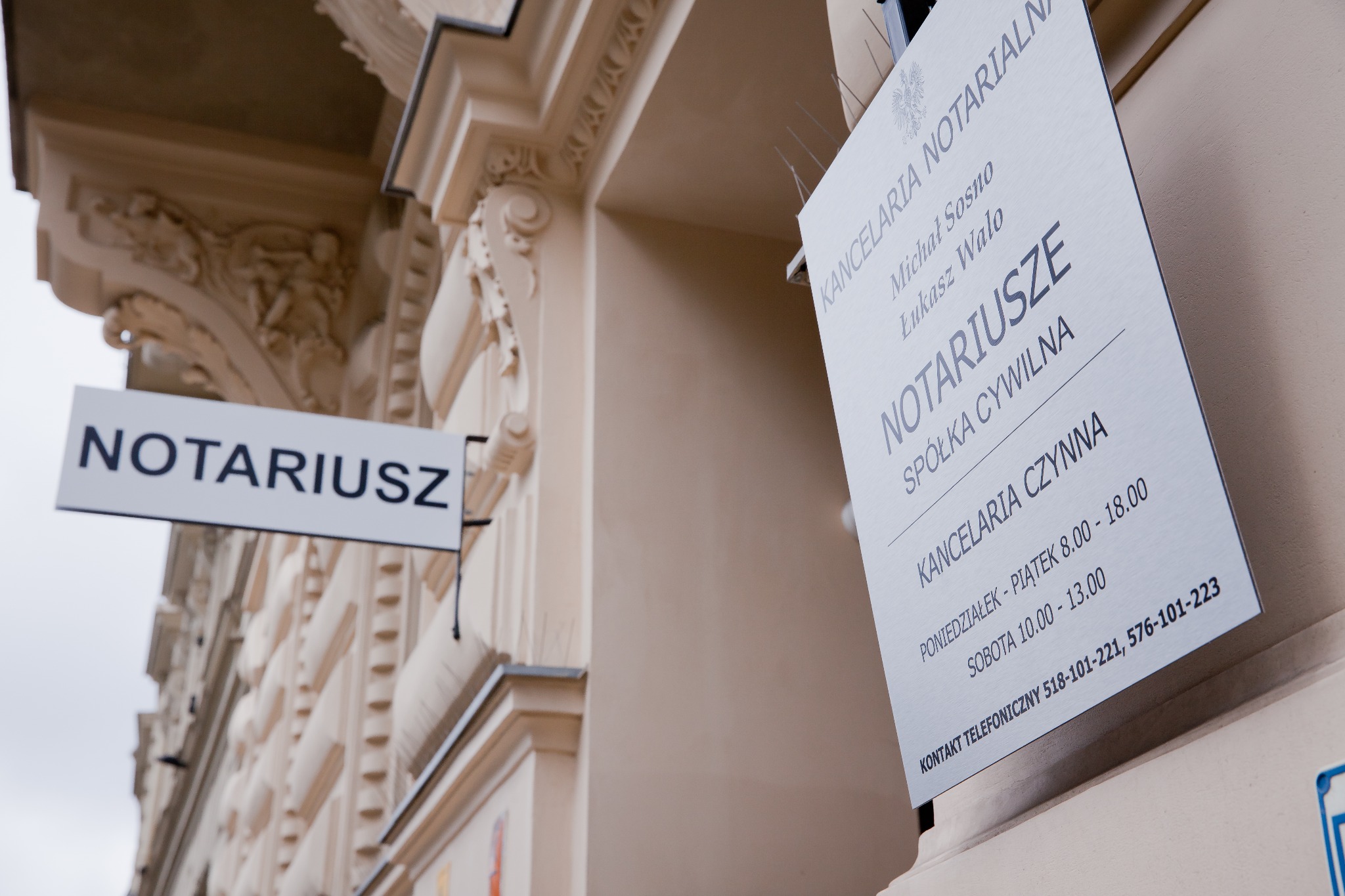 Zewnętrzny widok na budynek kancelarii z dwoma szyldami informacyjnymi: jeden z napisem 'Notariusz', drugi z danymi kontaktowymi, godzinami otwarcia i nazwiskami notariuszy.