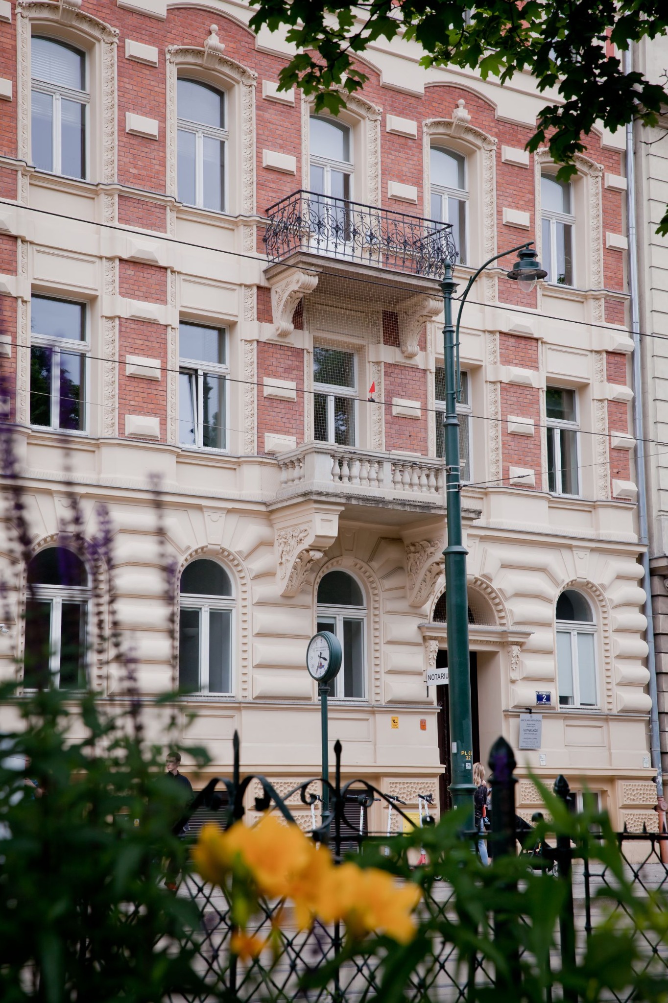 Fasada kamienicy z balkonami i szyldem notarialnym, widok przez ozdobne ogrodzenie z kwiatami.