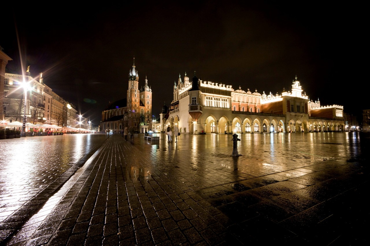 Nocny widok na Rynek Główny w Krakowie po deszczu, z Sukiennicami i Bazyliką Mariacką oświetlonymi sztucznym światłem, odbijającymi się w mokrej nawierzchni bruku.