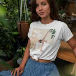 Kobieta w białej koszulce z grafiką jogi 'Self Care First' na tle zieleni. Koszulka zawiązana w supeł, niebieska spódnica. Uśmiechnięta, naturalny makijaż.