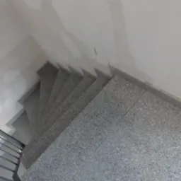 Wykończone schody wewnętrzne wyłożone szarym, nakrapianym gresem, widoczne z góry, z fragmentem metalowej barierki po lewej stronie i surową ścianą w tle.