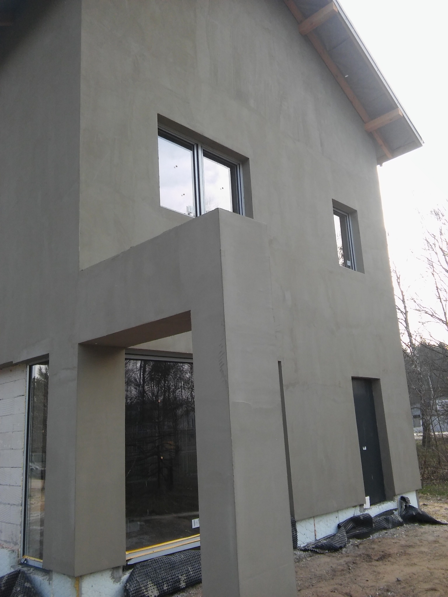 Surowy, szary budynek z oknami i drzwiami, w trakcie prac wykończeniowych elewacji, widoczne bloczki z betonu komórkowego i folia fundamentowa.