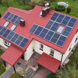 Dom jednorodzinny z czerwoną dachówką, na której zamontowane są panele słoneczne; widok z góry, z anteną satelitarną na ścianie i basenem w ogrodzie.