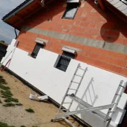 Ściana domu w trakcie ocieplania styropianem, widoczne rusztowanie, cegła dziurawka i okna z roletami zewnętrznymi.