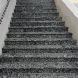 Schody zewnętrzne wyłożone ciemnym, nakrapianym kamieniem, z szarą balustradą i fragmentem fasady budynku w tle.