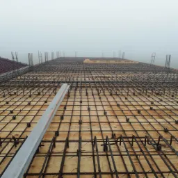 Siatka zbrojeniowa ułożona na drewnianym szalunku, widoczny poziomica, w tle ceglany komin i mgła.