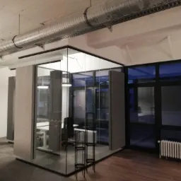 Nowoczesne biuro z przeszklonymi ścianami działowymi, widoczne biurka, krzesła i system wentylacyjny pod sufitem. Częściowo widoczne kartony sugerują prace wykończeniowe.