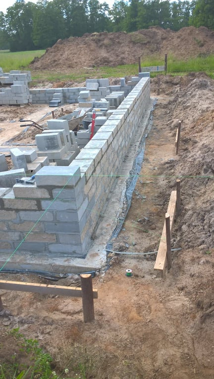 Wznoszenie muru fundamentowego z bloczków betonowych, widoczne sznurki poziomujące, przygotowane bloczki, wykop i izolacja przeciwwilgociowa.