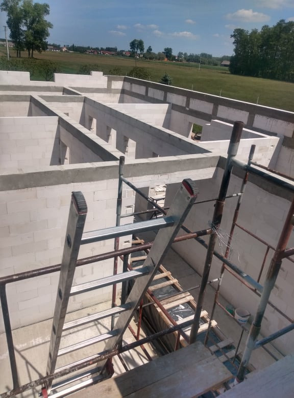Wnętrze budynku w trakcie budowy z białych bloczków, widoczne rusztowanie i drabina, w tle zielone pola i drzewa.