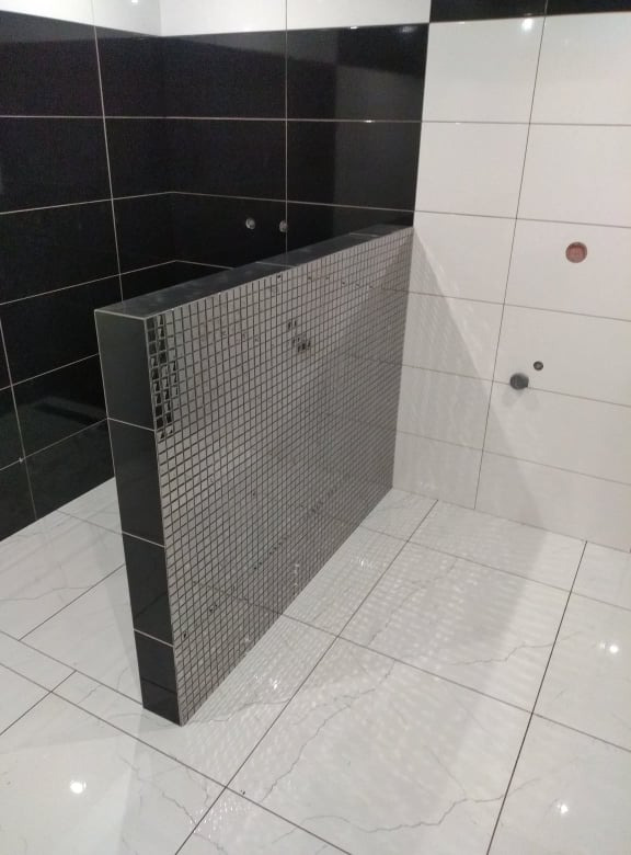 Wnętrze łazienki w trakcie prac wykończeniowych; widoczna ścianka działowa z mozaiki w kolorze szarym, czarno-białe płytki na ścianach oraz biała marmurowa podłoga.